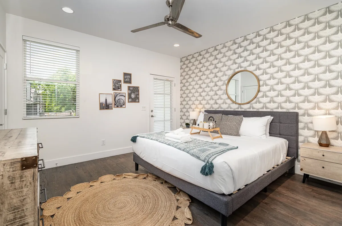 A spacious bedroom featuring a large king-sized bed with a decorative throw, geometric wallpaper, a round mirror, and wall art. Natural light streams in from the window, creating a serene and cozy ambiance ideal for relaxation.