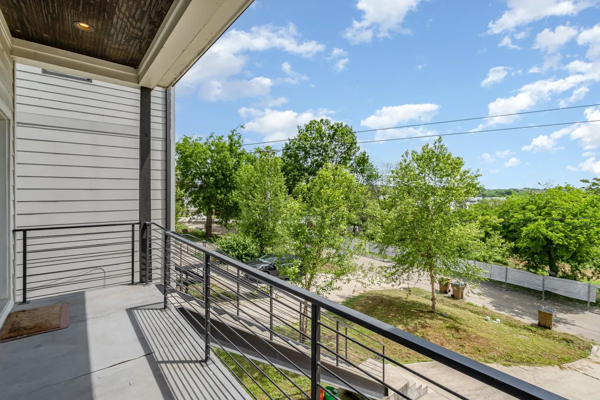 A private balcony with a sleek railing overlooks green trees and a blue sky. The outdoor space invites relaxation, with room to enjoy morning coffee, read a book, or watch the neighborhood in a tranquil setting.