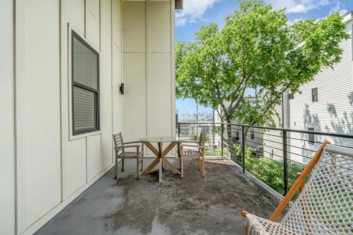 A tranquil patio scene features a table and chairs set beneath a leafy green tree, with a view of the city beyond. The space is designed for outdoor relaxation, meals, or reading, enhanced by the calming atmosphere.