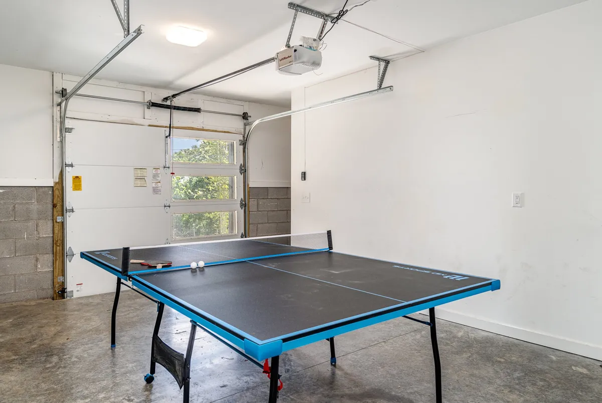 A brightly lit garage features a ping pong table at its center, with a large garage door and a window revealing an outdoor view. The space feels playful and relaxed, ideal for friendly competitions and casual gatherings.