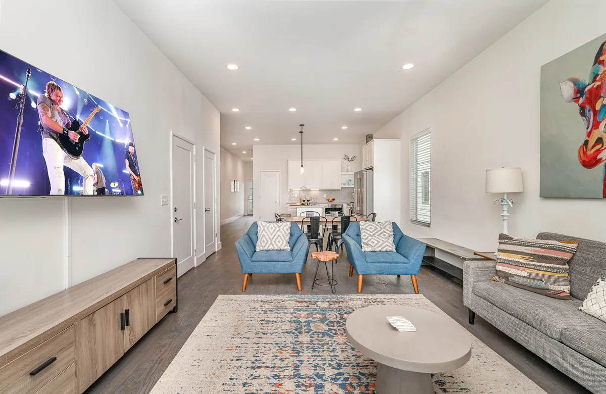 The image showcases a stylish, lively living room with plush blue accent chairs, a spacious coffee table, and a large television displaying a concert scene, all set in a bright, open space perfect for socializing or unwinding.