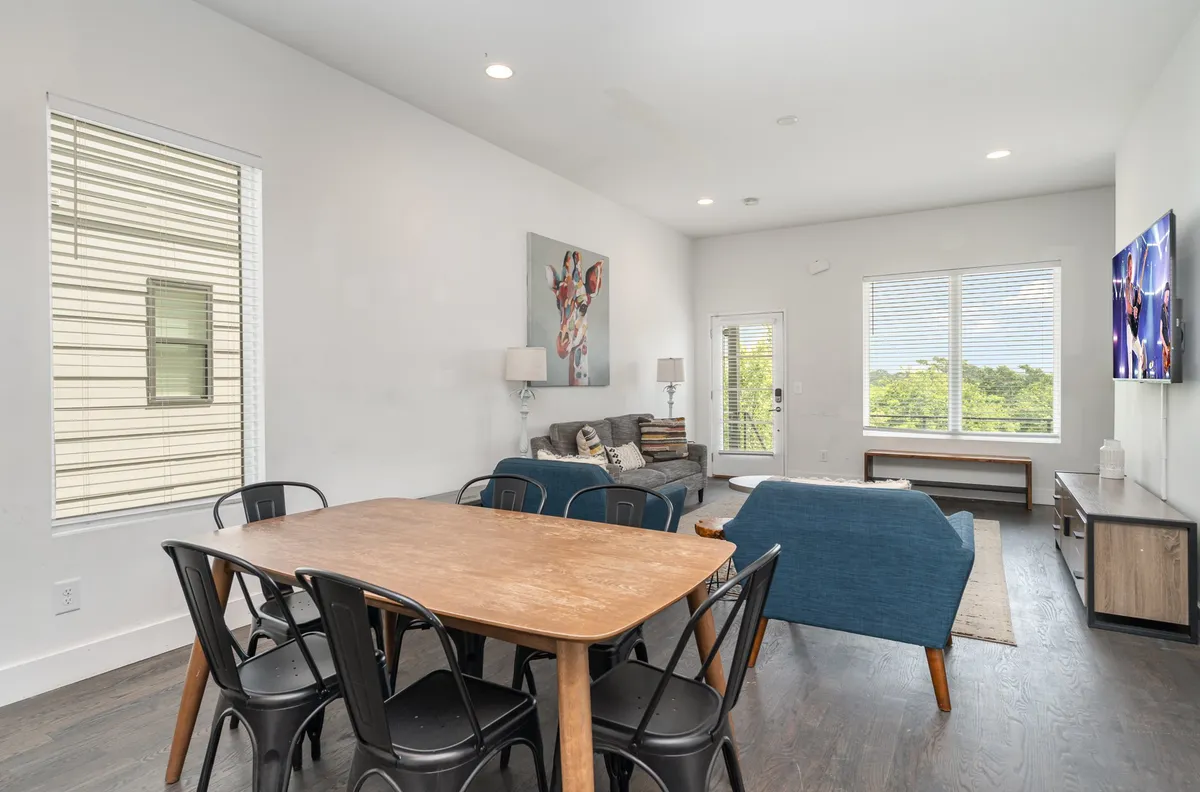 A spacious, open-plan living and dining area filled with natural light. The scene features a large dining table set for a group, blue sofas arranged for conversation or TV viewing, and contemporary artwork on the walls. Large windows let in ample daylight, creating a warm and welcoming atmosphere ideal for gatherings.